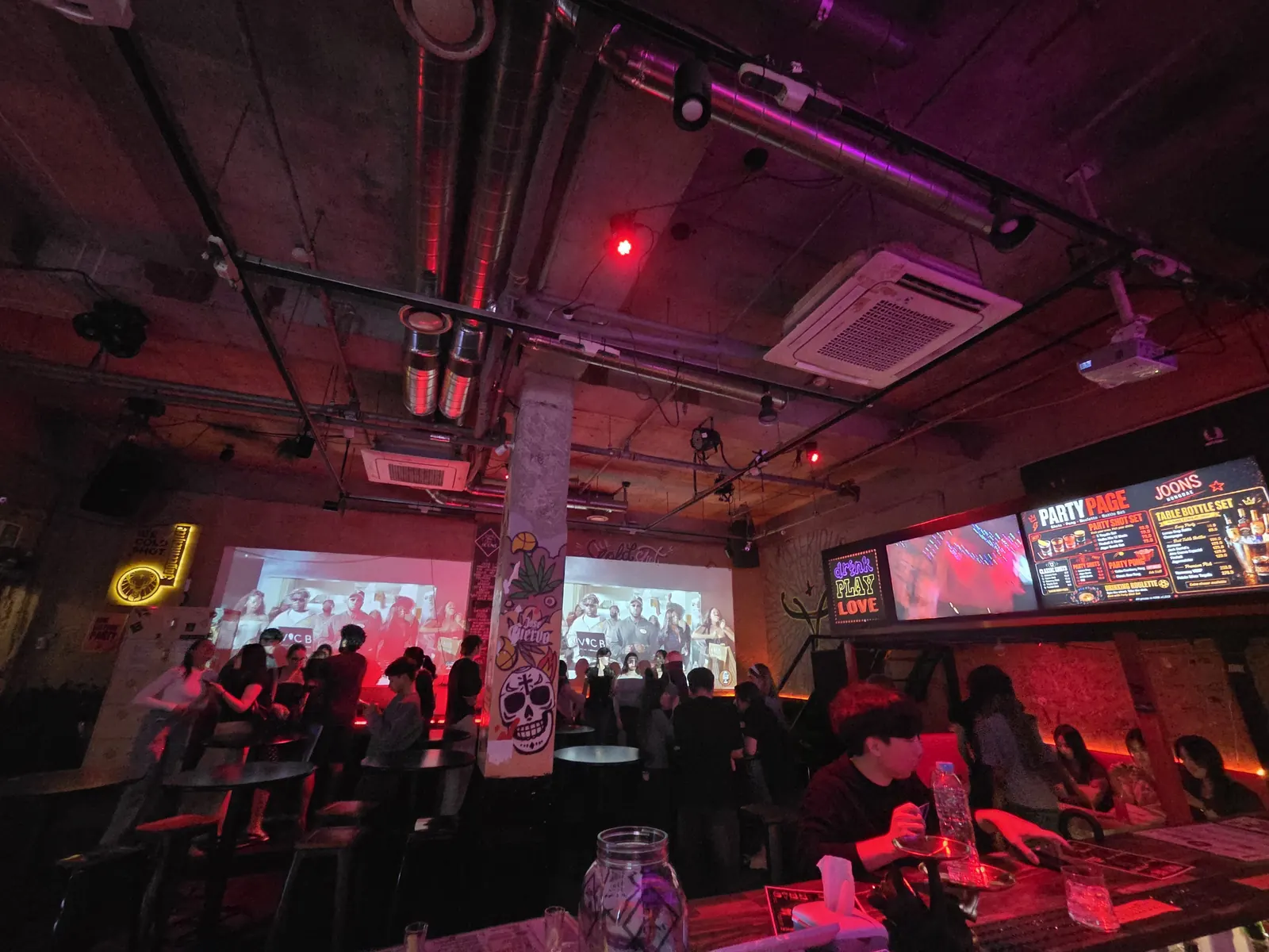 The main room at Joons, lit red, with projection screens and a standing crowd